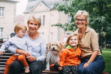 Zwei ältere Damen sitzen gemeinsam mit ihren Enkelkindern und einem Hund auf einer Parkbank und schauen lächelnd in die Kamera.