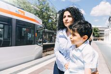 Eine Frau wartet mit ihrem Kind an einer Haltestelle, im Hintergrund fährt eine StadtBahn.