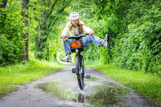 Eine Frau fährt mit einem Fahrrad von meinSiggi durch eine Pfütze und hebt dabei freudig die Beine an.