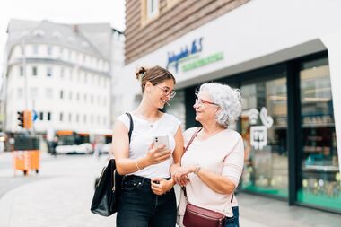 Zwei Frauen verschiedenen Alters stehen fröhlich vor dem Kundenzentrum Jahnplatz Nr. 5 und blicken sich an. Eine Frau hält ein Smartphone in der Hand.