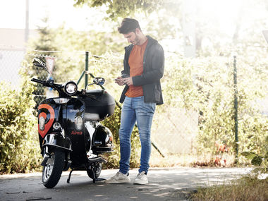 flowBie Sharing App runterladen, Roller buchen und los geht's! Ein Mann steht neben dem Alma E-Roller und schaltet den Roller mit seinem Handy frei.