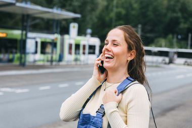 Junge Frau mit Blaumannhose steht telefonierend und lachend an einer Haltestelle.