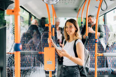Eine Frau steht mit ihrem Smartphone fr&ouml;hlich im Bus und f&auml;hrt mit eezy.nrw einfach und g&uuml;nstig durch ganz NRW.
