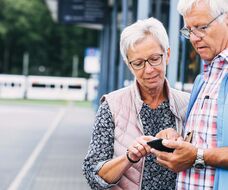 Ehepaar schaut an einer Haltestelle aufs Handy, im Hintergrund f&auml;hrt eine Vamos-StadtBahn.