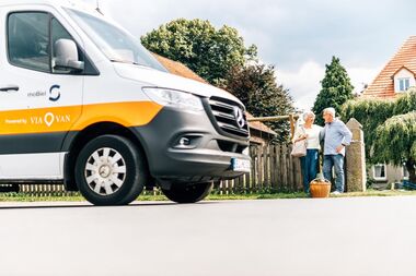 meinAnton fährt dich fast von Tür zu Tür. meinAnton-Fahrzeug mit zwei Personen im Hintergrund, die vor einem Haus warten.