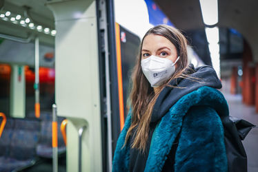 Eine Frau mit FFP2-Maske wartet in einer StadtBahn-Haltestelle. Im Hintergrund ist eine Vamos-Bahn zu sehen.