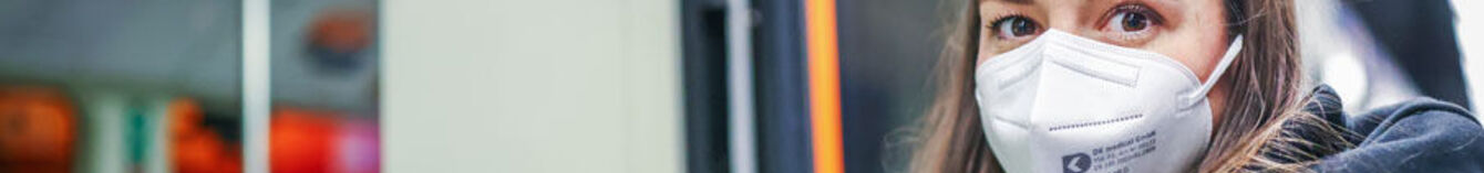 Eine Frau mit FFP2-Maske wartet in einer StadtBahn-Haltestelle. Im Hintergrund ist eine Vamos-Bahn zu sehen.