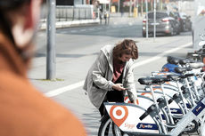 Eine Frau entsperrt im Hintergrund ein Siggi-Fahrrad zur Ausleihe, im Vordergrund ist ein Mann mit Fahrradhelm zu sehen