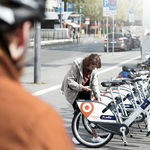 Eine Frau entsperrt im Hintergrund ein Siggi-Fahrrad zur Ausleihe, im Vordergrund ist ein Mann mit Fahrradhelm zu sehen