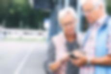 Ehepaar schaut an einer Haltestelle aufs Handy, im Hintergrund fährt eine Vamos-StadtBahn.