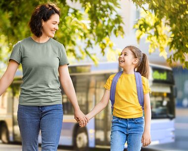 Frau und Kind gehen Hand in Hand fr&ouml;hlich einen Weg entlang. Im Hintergrund ist ein Bus zu sehen. | Mit dem kostenlosen ElternKindTicket den Schulweg &uuml;ben.