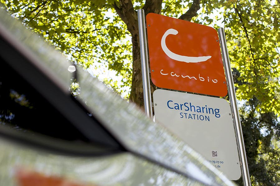 Leihen Sie Ihr cambio-Auto an einer der Stationen in Haltestellen-Nähe. Detailaufnahme des cambio-Schilds an einer cambio-Station und ein unscharfer Ausschnitt eines Autos davor.